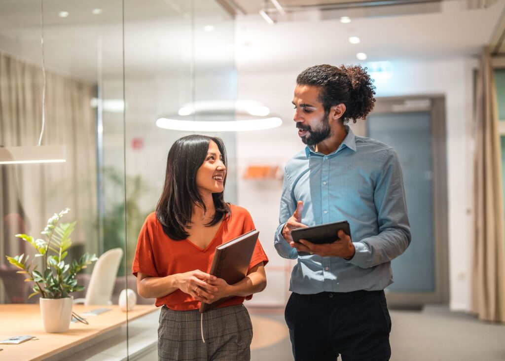 Two colleagues, a man and a woman, walk and talk in a modern office, discussing a tablet.