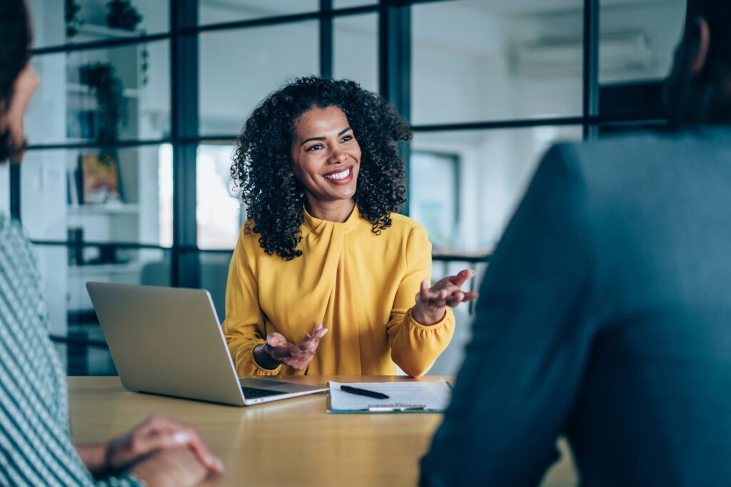 Businesswoman in yellow shirt talks to colleagues at a table with a laptop.