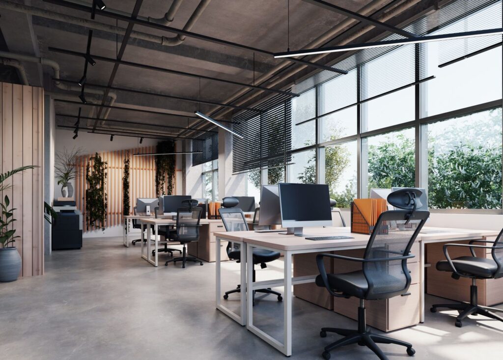 Modern open-plan office with desks, chairs, monitors, and large windows overlooking greenery.